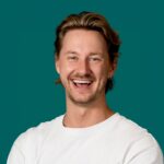 Vrolijke zakelijke profielfoto van een lachende man die menselijkheid en eigenheid uitstraalt tegen een groene achtergrond.